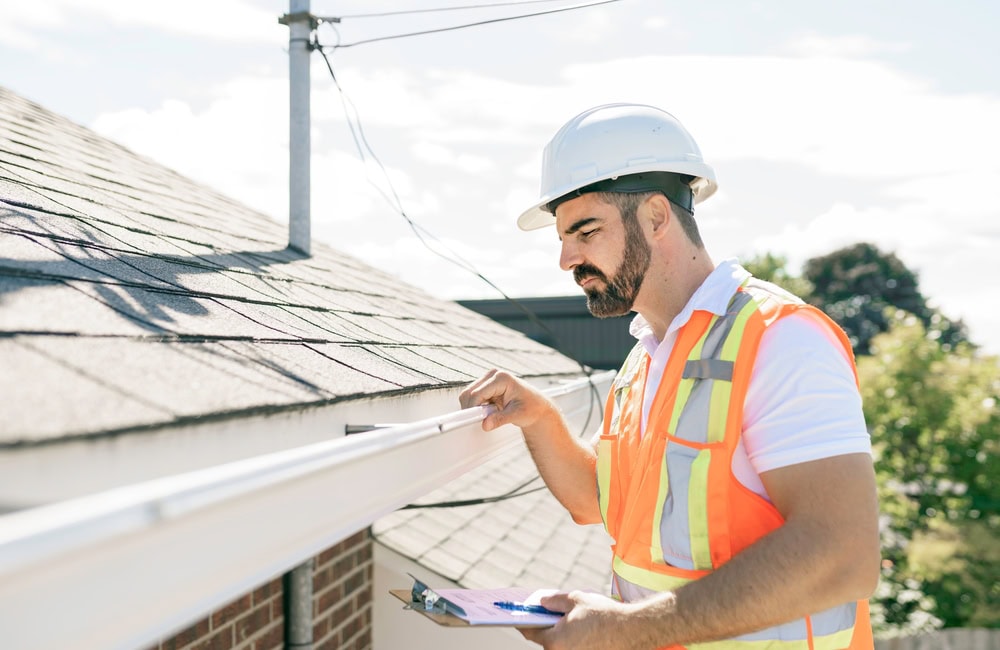 A construction worker in a hard hat and safety vest inspects a house gutter with a clipboard, standing beside a roof under bright daylight—perfect for any home improvement Suffolk County project or roofing contractor Suffolk County inspection.