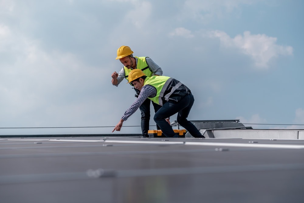 Two workers in yellow safety helmets and reflective vests examine solar panels on a rooftop. As part of a trusted roofing contractor Suffolk County, NY team, they focus intently on the inspection for quality home improvement results.