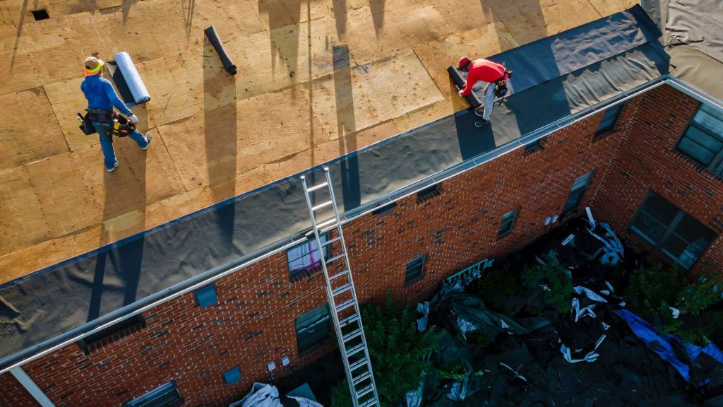 Aerial view of two workers on a flat roof, one laying material and the other carrying tools. A ladder leans against the brick building, highlighting a roofing contractor Suffolk County, NY home improvement project below.