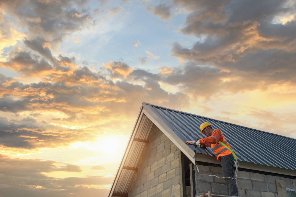 A construction worker in a safety vest and helmet installs metal roofing on a building at sunset in NY, with dramatic clouds and sunlight—showcasing the skills of a trusted roofing contractor Suffolk County.