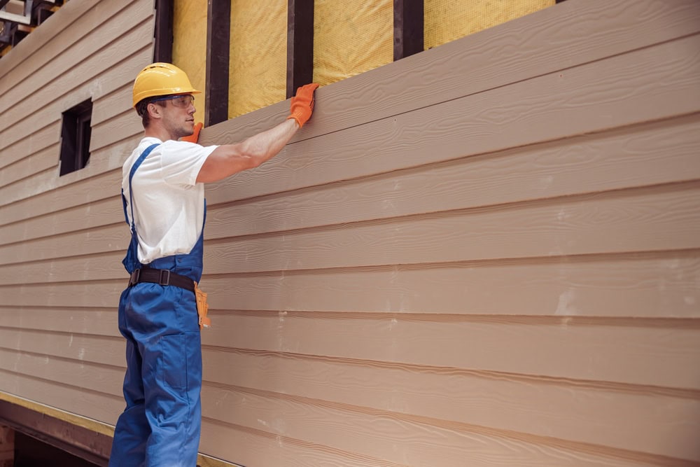 A construction worker in blue overalls, a yellow hard hat, and orange gloves installs beige siding panels on the exterior wall of a building under construction in NY for a home improvement Suffolk County project.