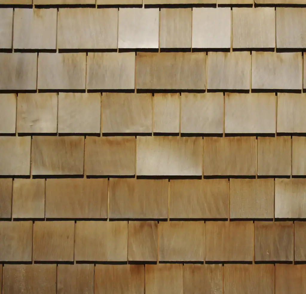 A close-up view of a wall covered with overlapping rectangular wooden shingles, arranged in horizontal rows, showing natural wood grain and varying light and dark brown tones.