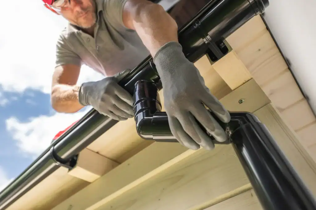 A person wearing gloves and glasses installs a black rain gutter system on the edge of a wooden roof under a bright sky, showcasing expert home improvement in Suffolk County, NY.