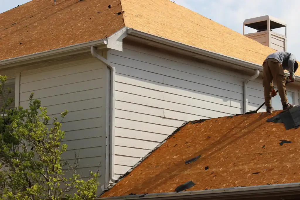 A person works on the roof of a house, replacing shingles. The roof is covered with wooden boards, and some old shingles and debris are scattered. The house is white with horizontal siding and a chimney visible.
