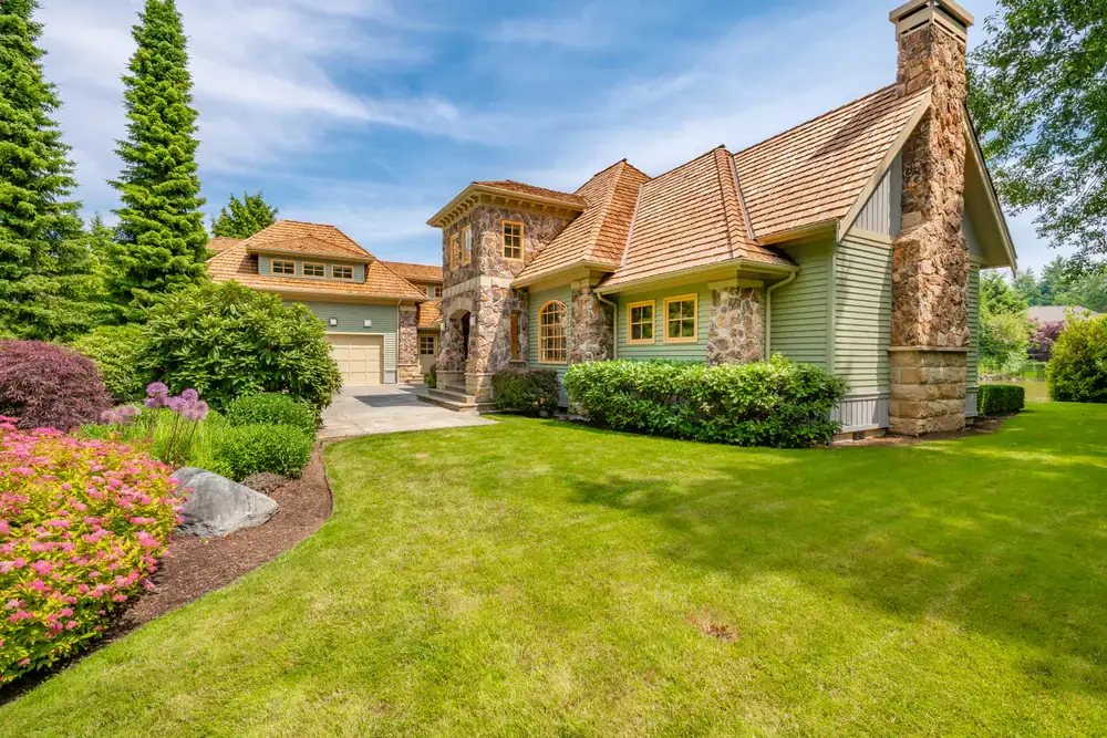 A large suburban house with stone and wood siding, multiple gables, a chimney, and well-manicured green lawn and garden, surrounded by trees and shrubs under a bright, blue sky.