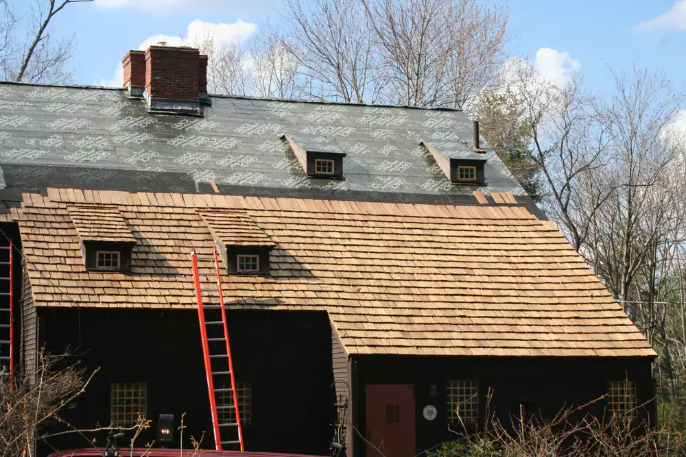 A wooden house with a partially completed roof; the lower half has new wooden shingles, while the upper half is covered in roofing underlayment. Two red ladders lean against the house under a blue sky with scattered clouds.