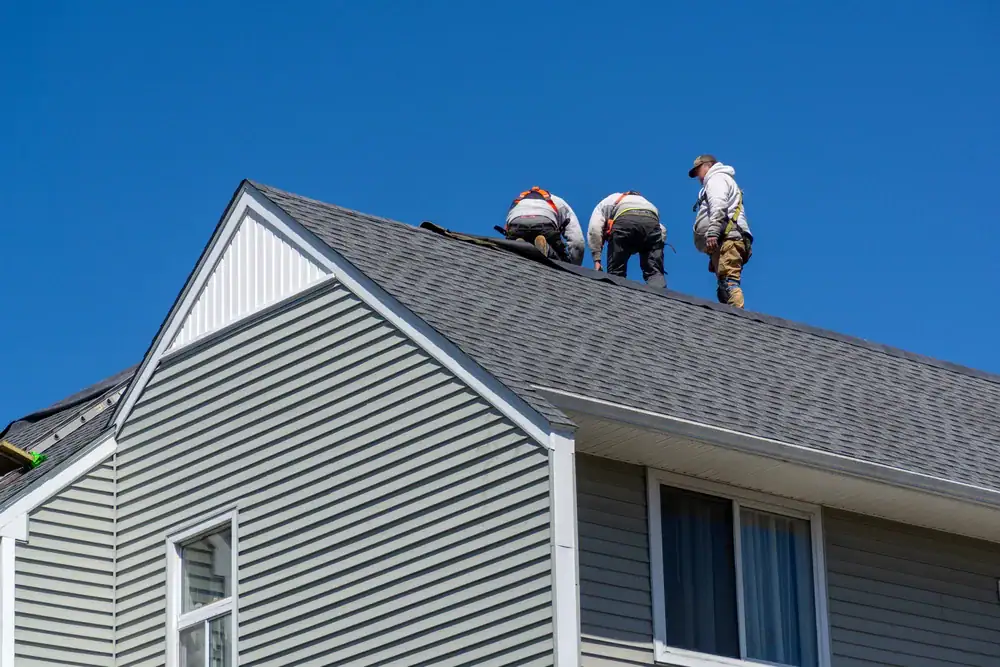 Three workers wearing protective gear are installing or repairing shingles on the roof of a light gray house with white trim, under a clear blue sky.