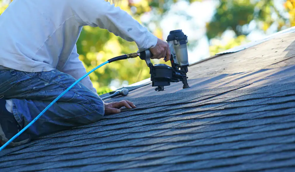 A person kneels on a roof, using a nail gun to secure asphalt shingles. The individual wears jeans and a long-sleeve shirt, working in daylight with trees visible in the blurred background.